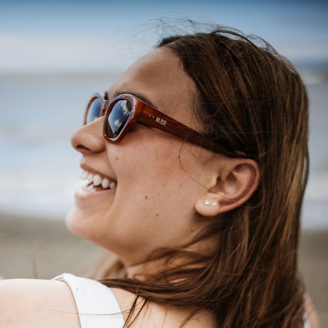 Close up of a womans face side on wearing burnt orange coloured plastic frame sunglasses with brown polarized lenses. A beach scene is out of focus in the background.