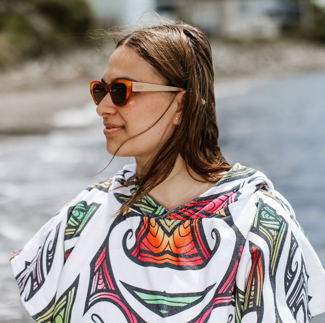 Woman at the beach wearing burnt orange coloured plastic frame sunglasses with brown polarized lenses and wooden arms. She also has a white towel hoodie on with multi coloured kory pattern.