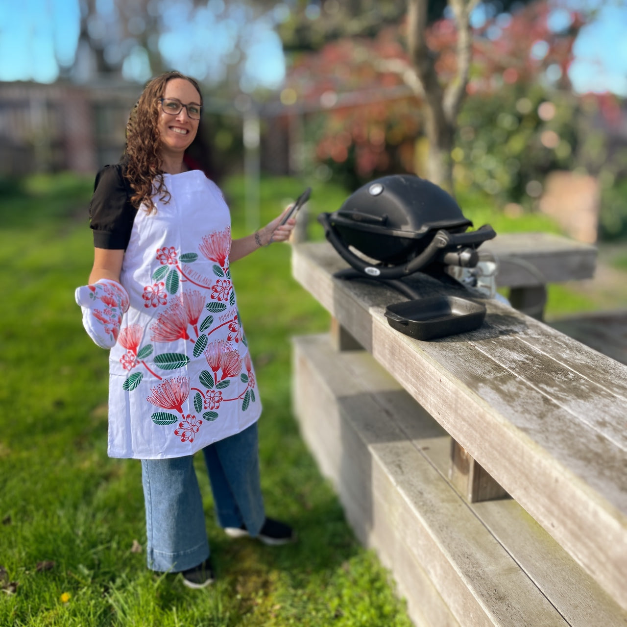Woman at bbq wearing white apron with red and green pohutukawa print with matching oven mitt.