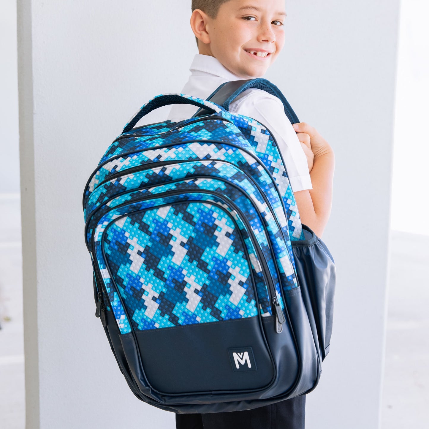 Young boy looking over his shoulder while holding onto a Childrens backpack by brand Montiico that is multi shades of blue with a block print on the top half and navy blue on the bottom.