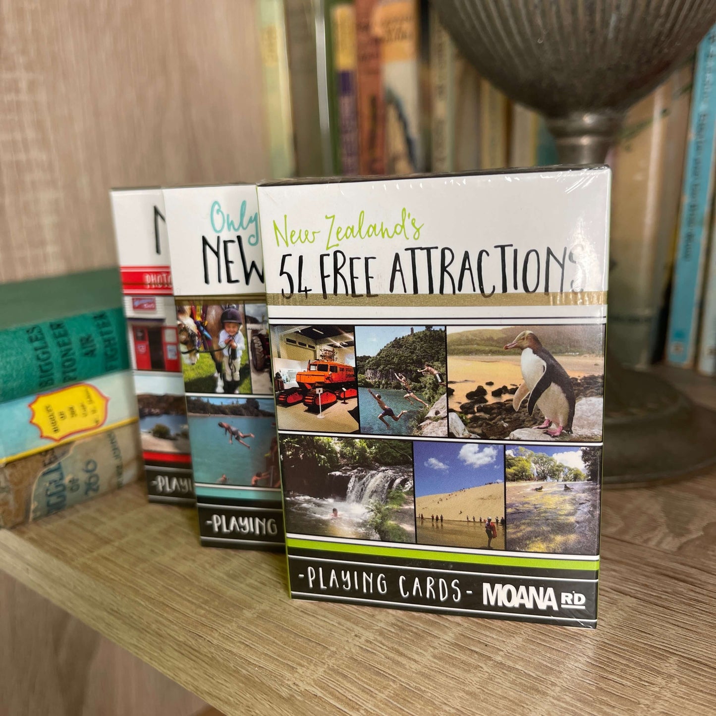 Pack of playing cards featuring NZ attractions sitting on a book shelf with other card packs behind it.