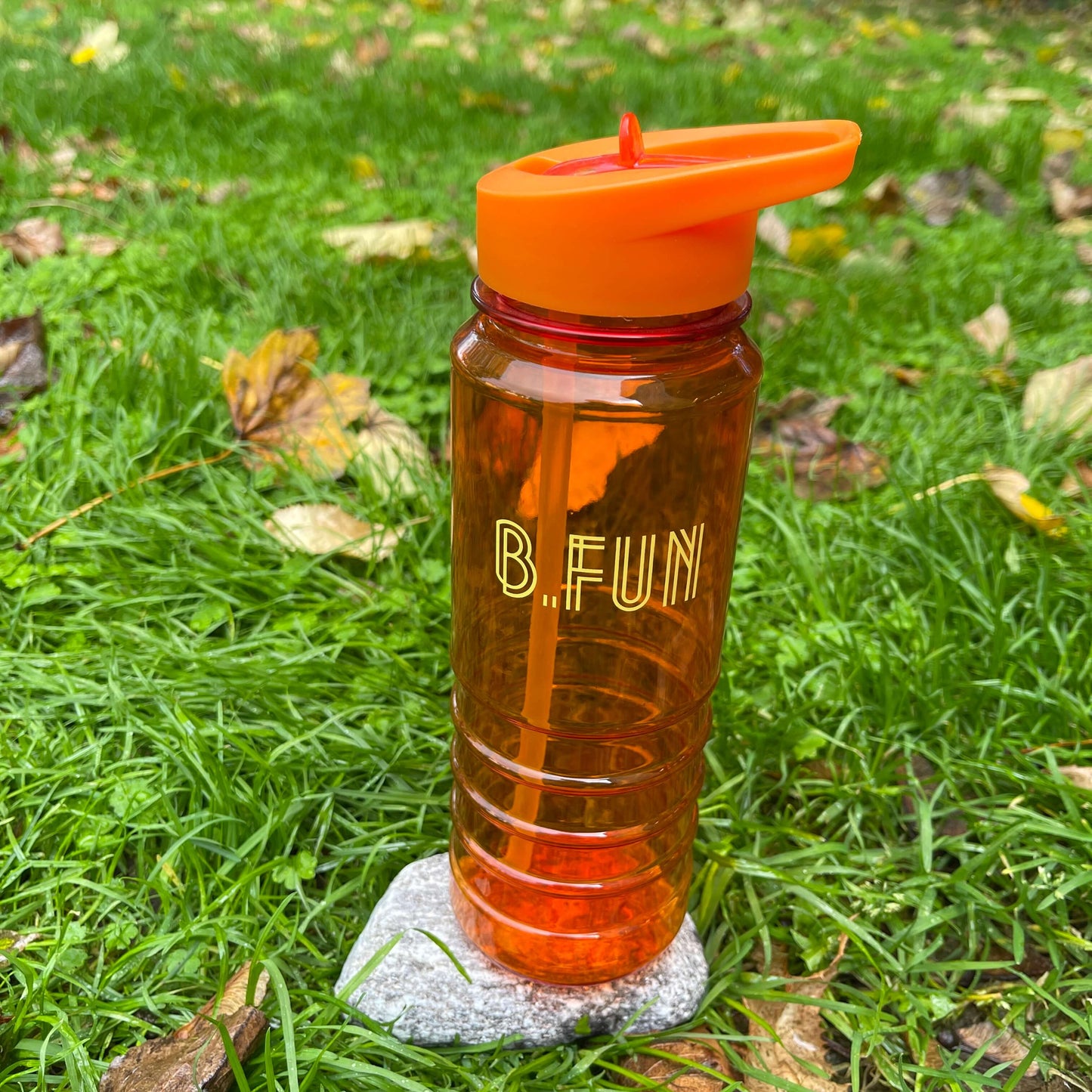 Bright orange plastic sports water bottle with B=Fun printed on it sitting on a rock in the grass.
