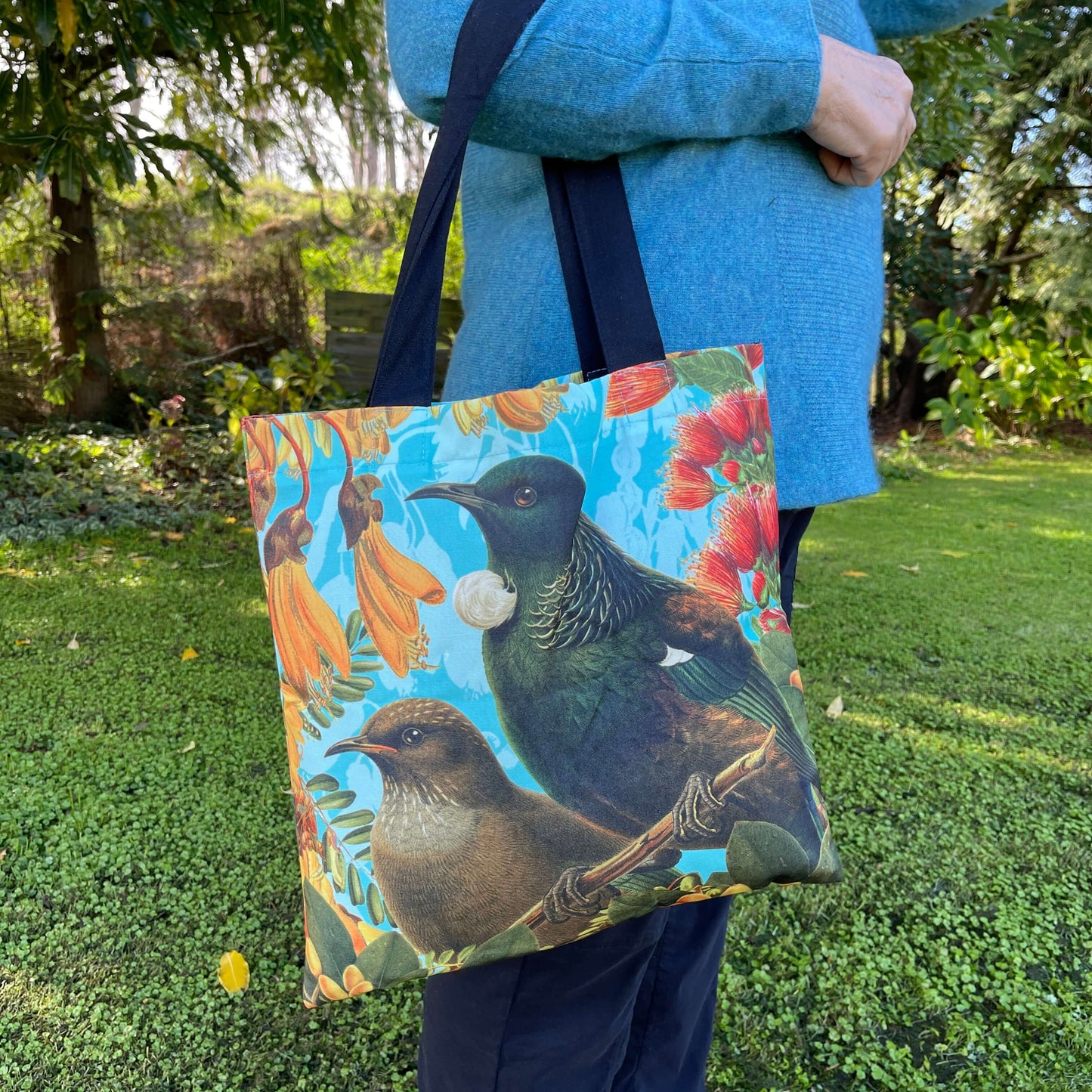 Woman with a tote bag over her arm. The tote features a Bellbird & Tui amongst Kowhai & Pohutukawa flowers.