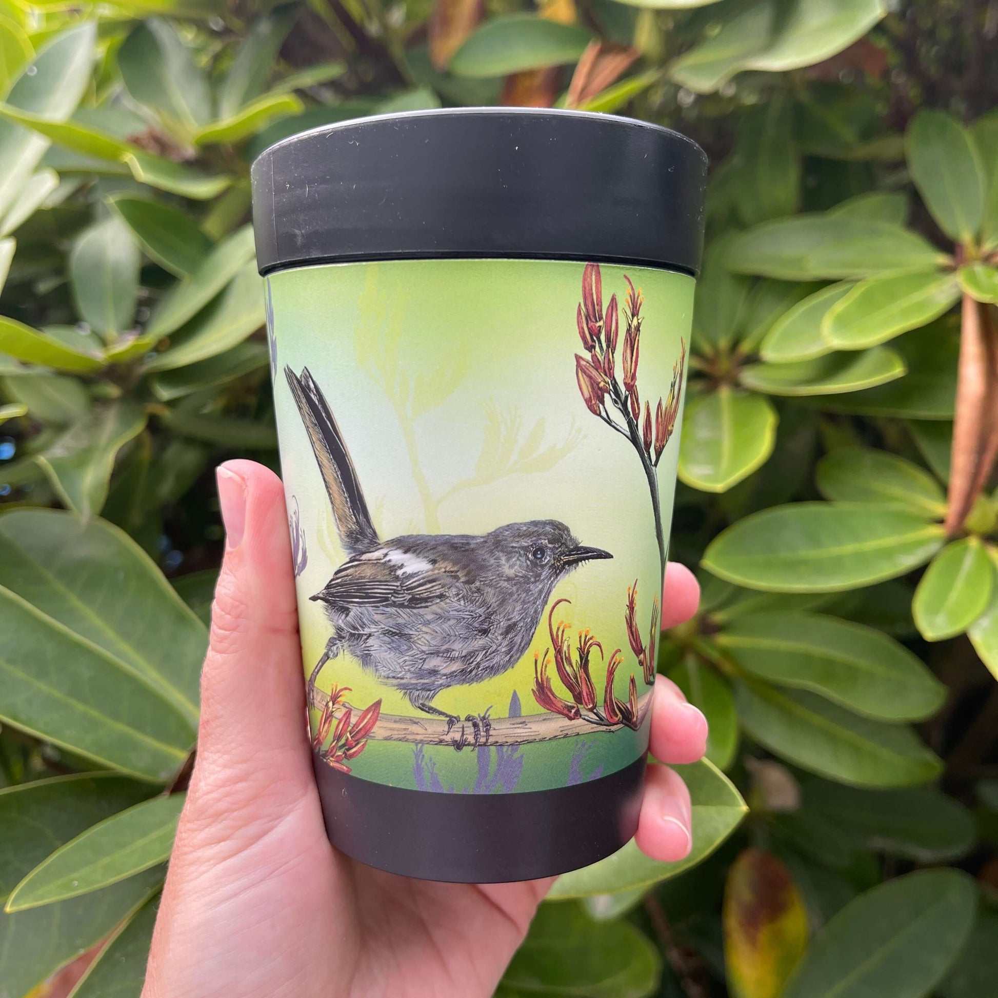 Black reusable coffee cup with Hihi birds and Flax flowers on a green background.