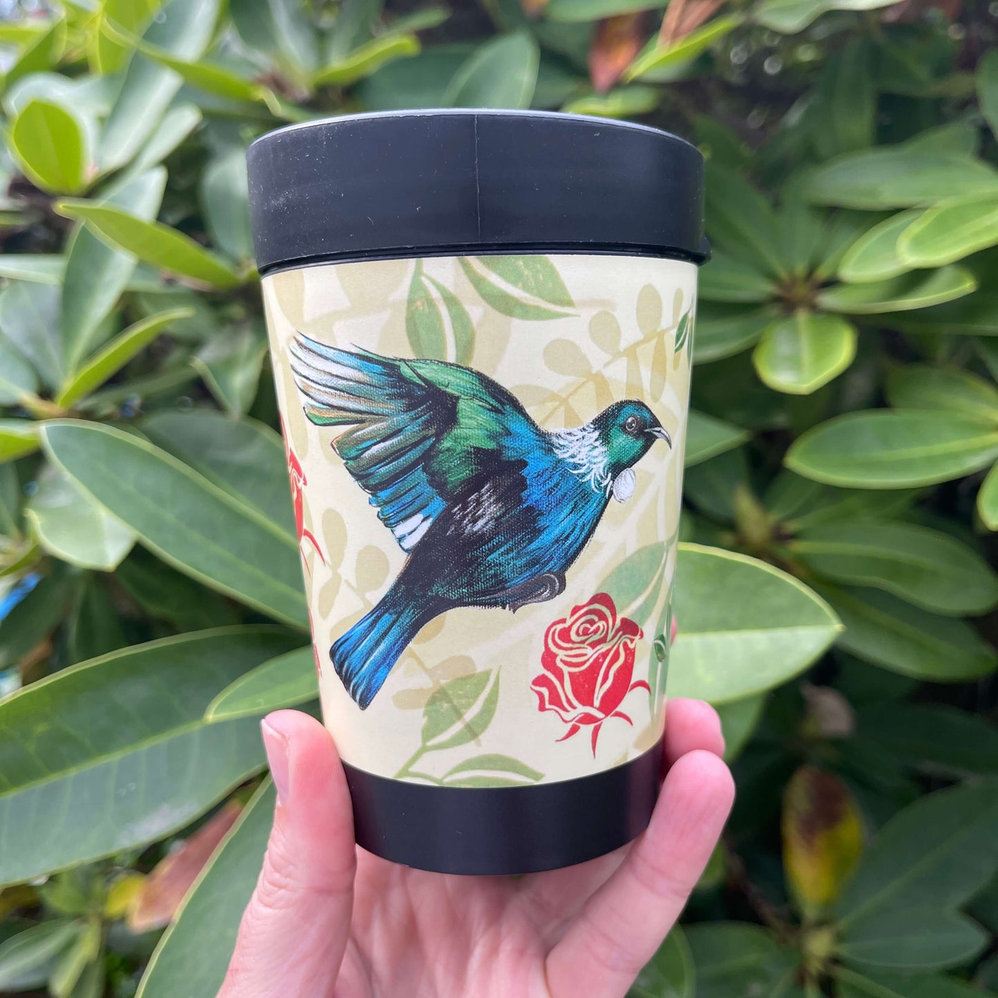 Black reusable coffee cup with beige and green leaf patterned wrap featuring the New Zealand Tui bird and red roses.