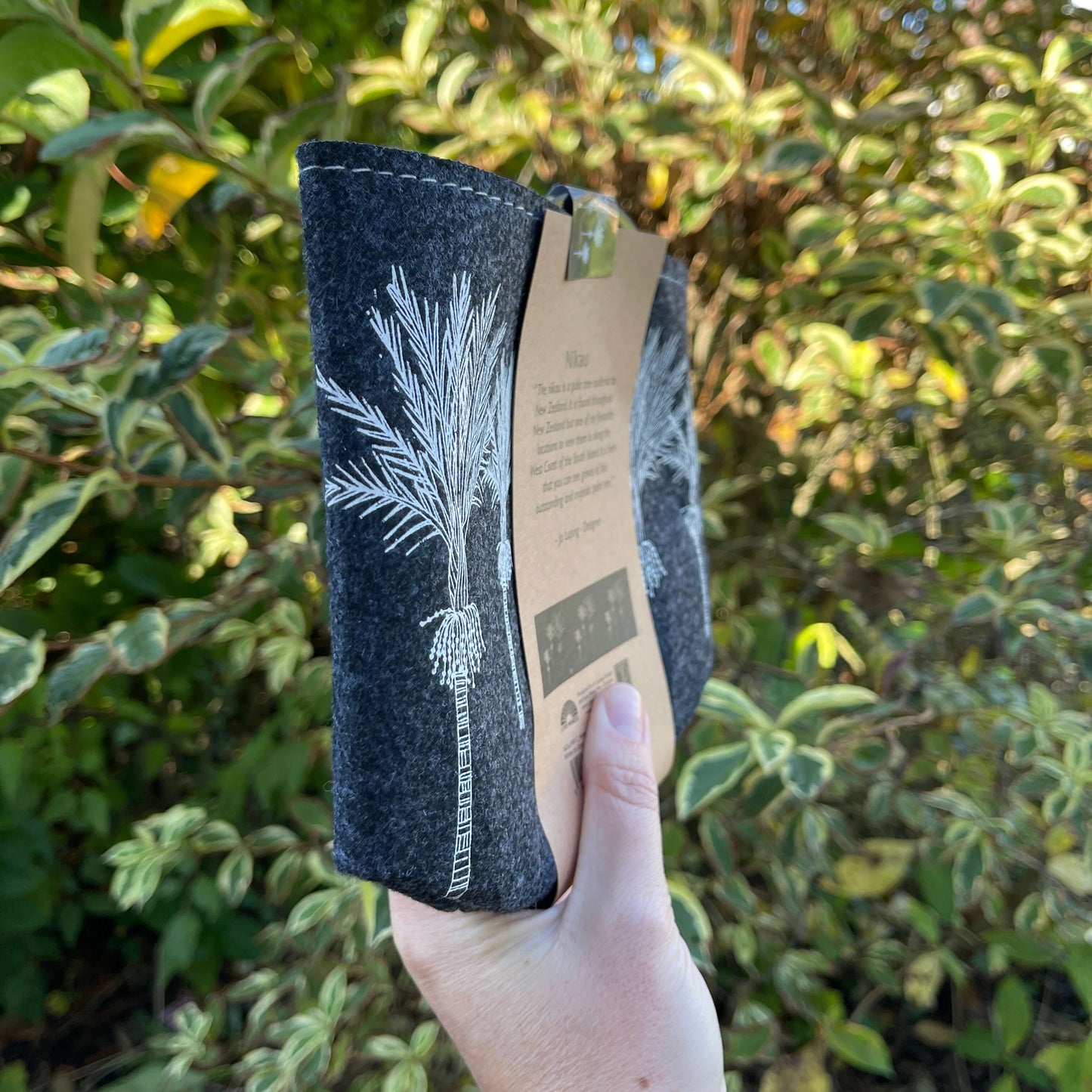 Dark grey felt planter with white Nikau trees printed on it sitting in a grey dish, flat packed and wrapped with kraft paper.