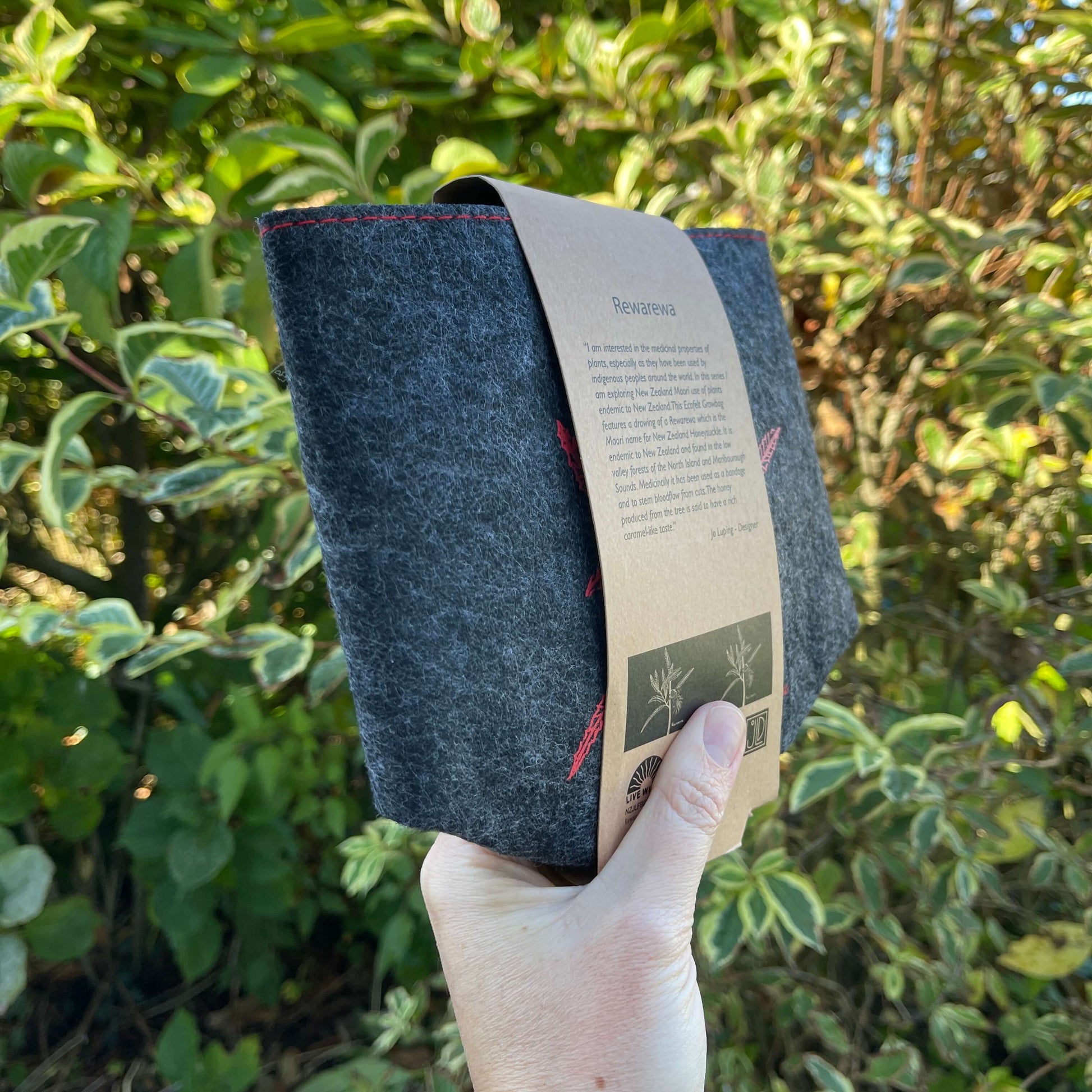 Dark grey felt planter with a rewarewa plant printed on it in red and sitting in a grey dish, flat packed and wrapped with kraft paper.