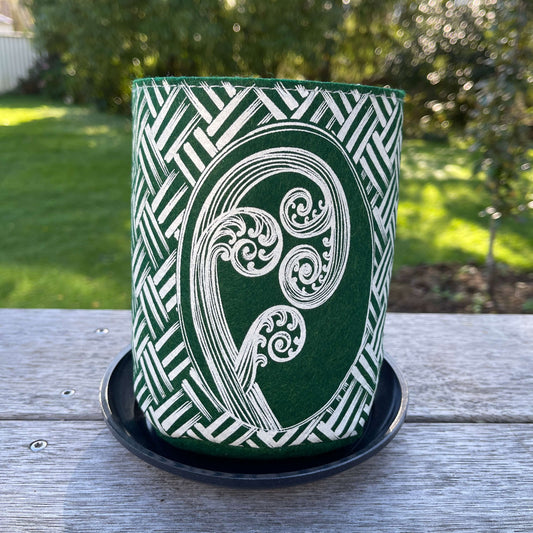 Green felt plant pot with white kete and ponga design, sitting in a dark grey saucer.