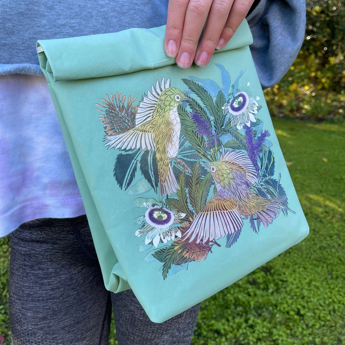Person holding a light blue folding paper style lunch bag with floral and bird design.
