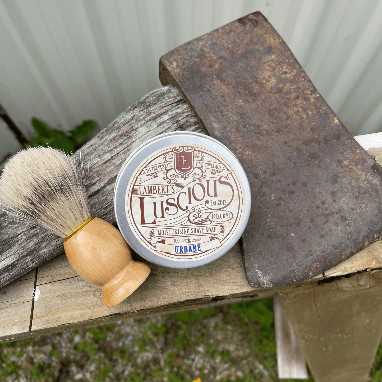 Metal tin of Lamberts Luscious mens shaving soap and a boar bristle shaving brush sitting next to an axe.