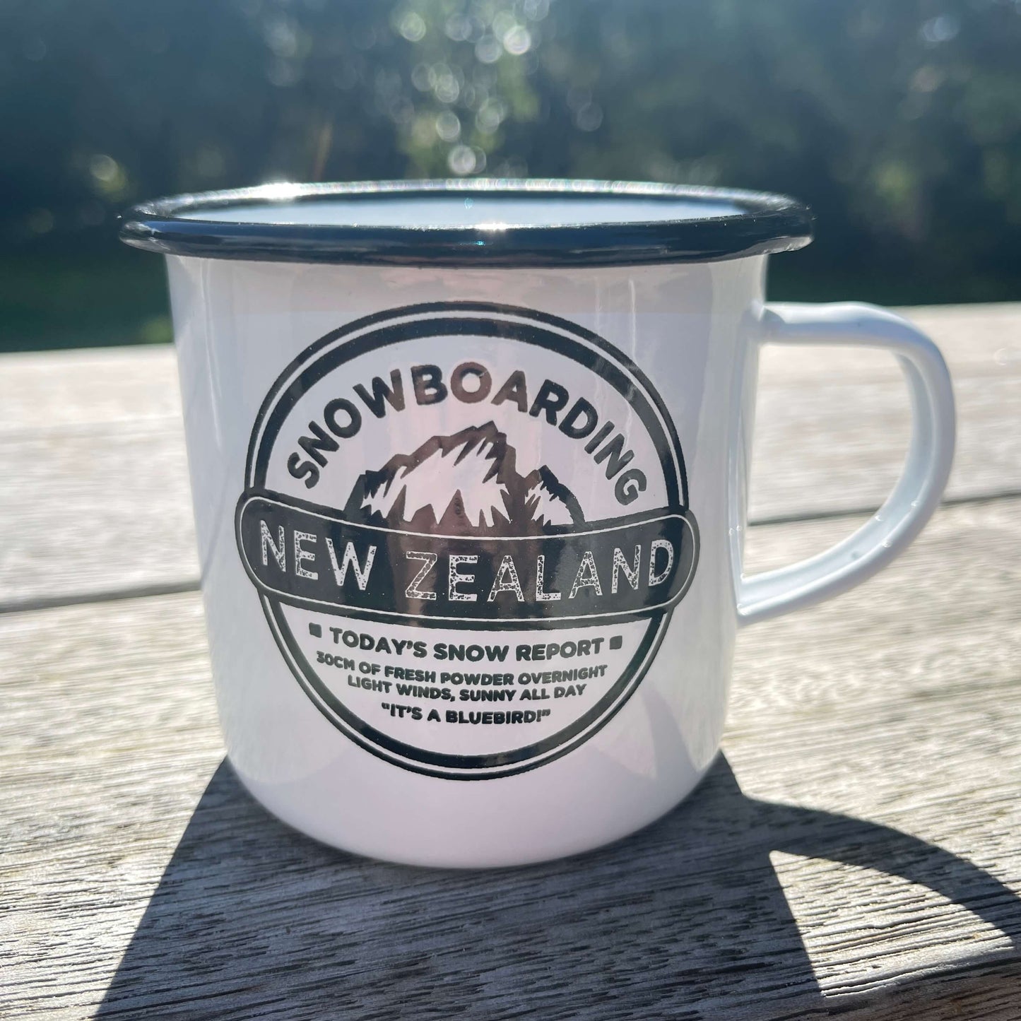 White enamel mug with black trim and image of a mountain with snowboarding new zealand written on it.