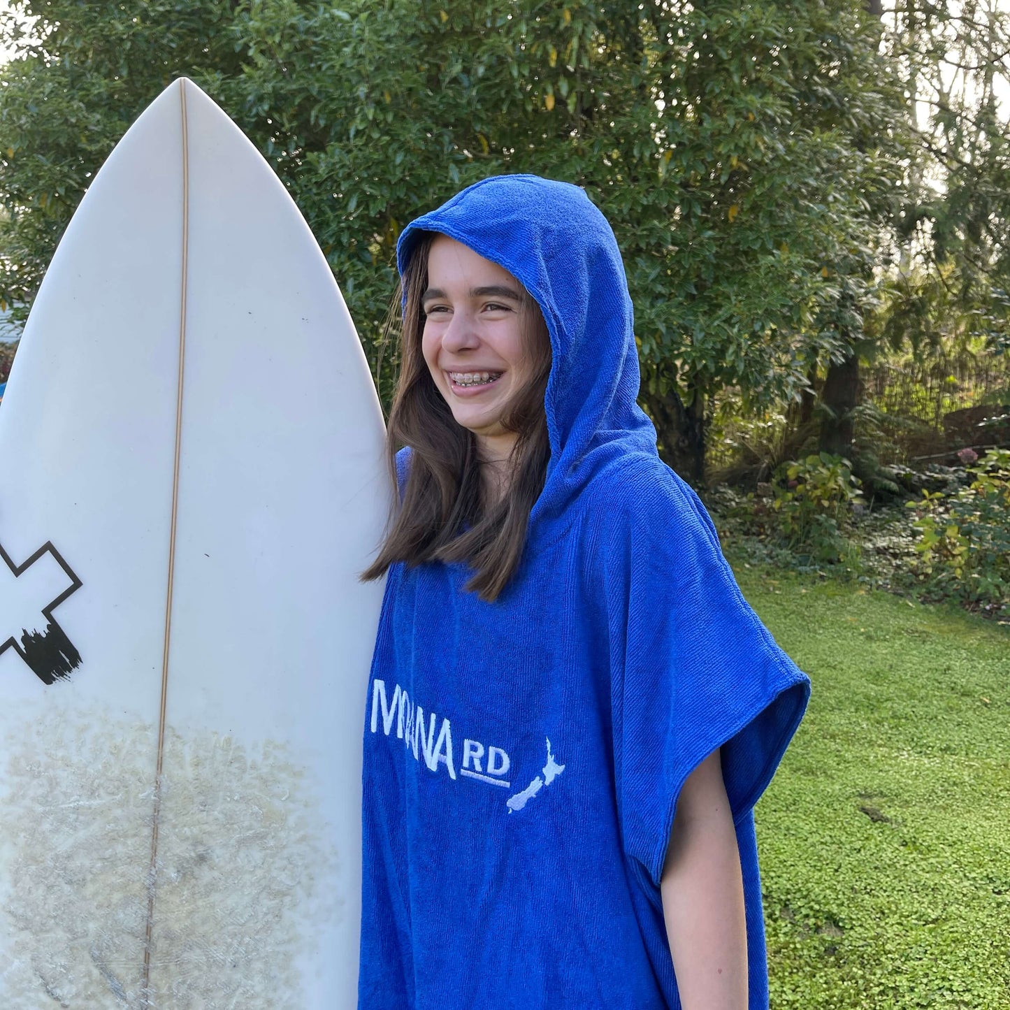 Young girl wearing a blue towel hoodie with the words Moana RD on it in white and a map of New Zealand. She is holding a surfboard.