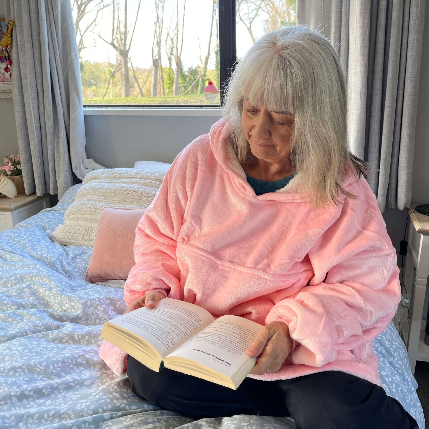 Woman sitting on a bed reading a book wearing an oversized pink hoodie.