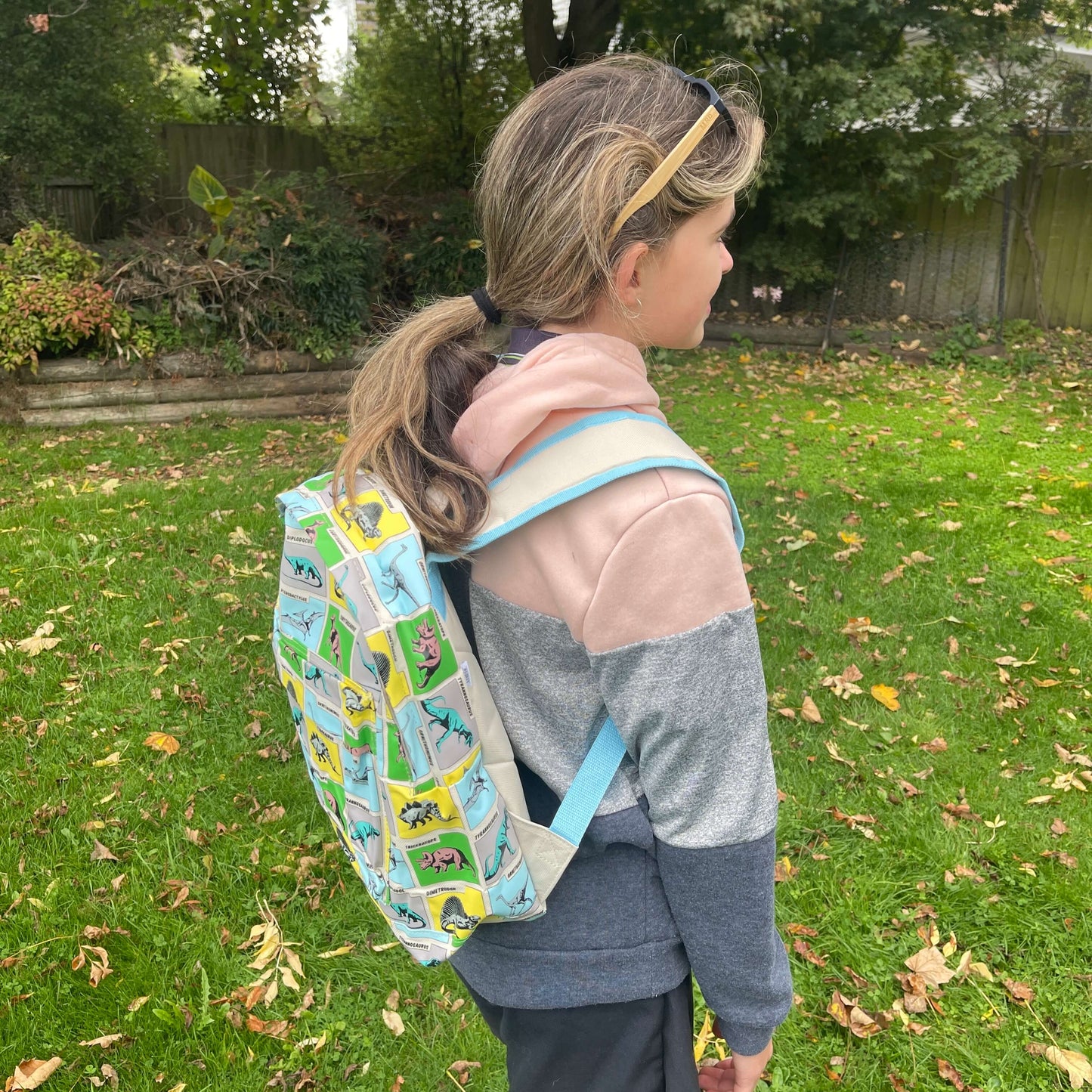 Girl wearing a backpack with dinosaurs printed on it in grey, blue, green & yellow squares.