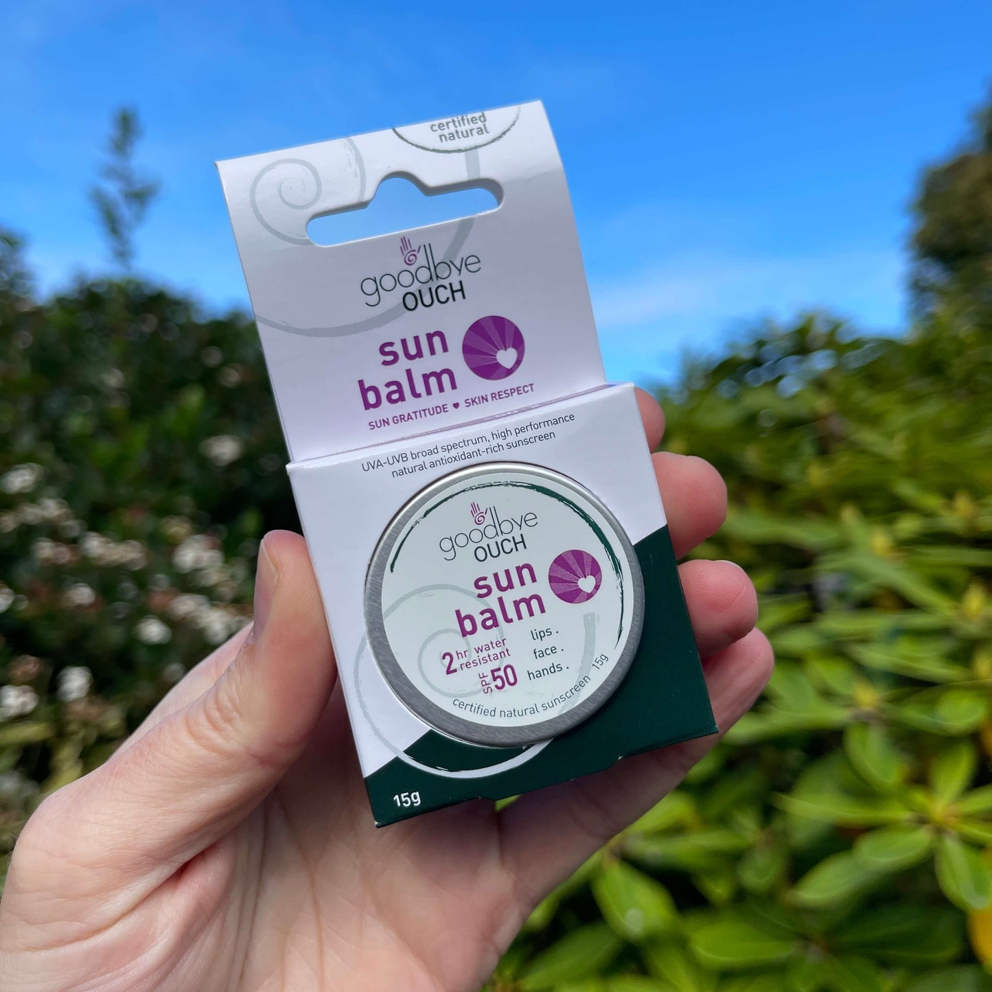 Womens hand holding a small sun balm tin in it's packaging with blue sky in the background.