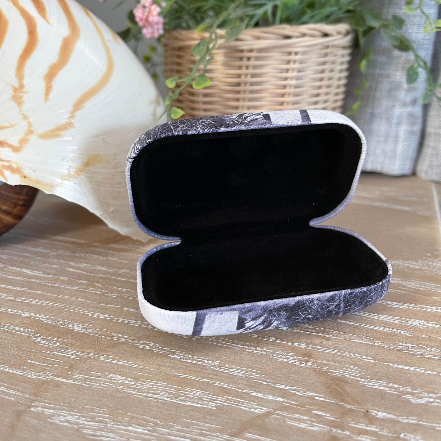 Small trinket case opened with black lining sitting on a table with a large shell and faux pot plant.