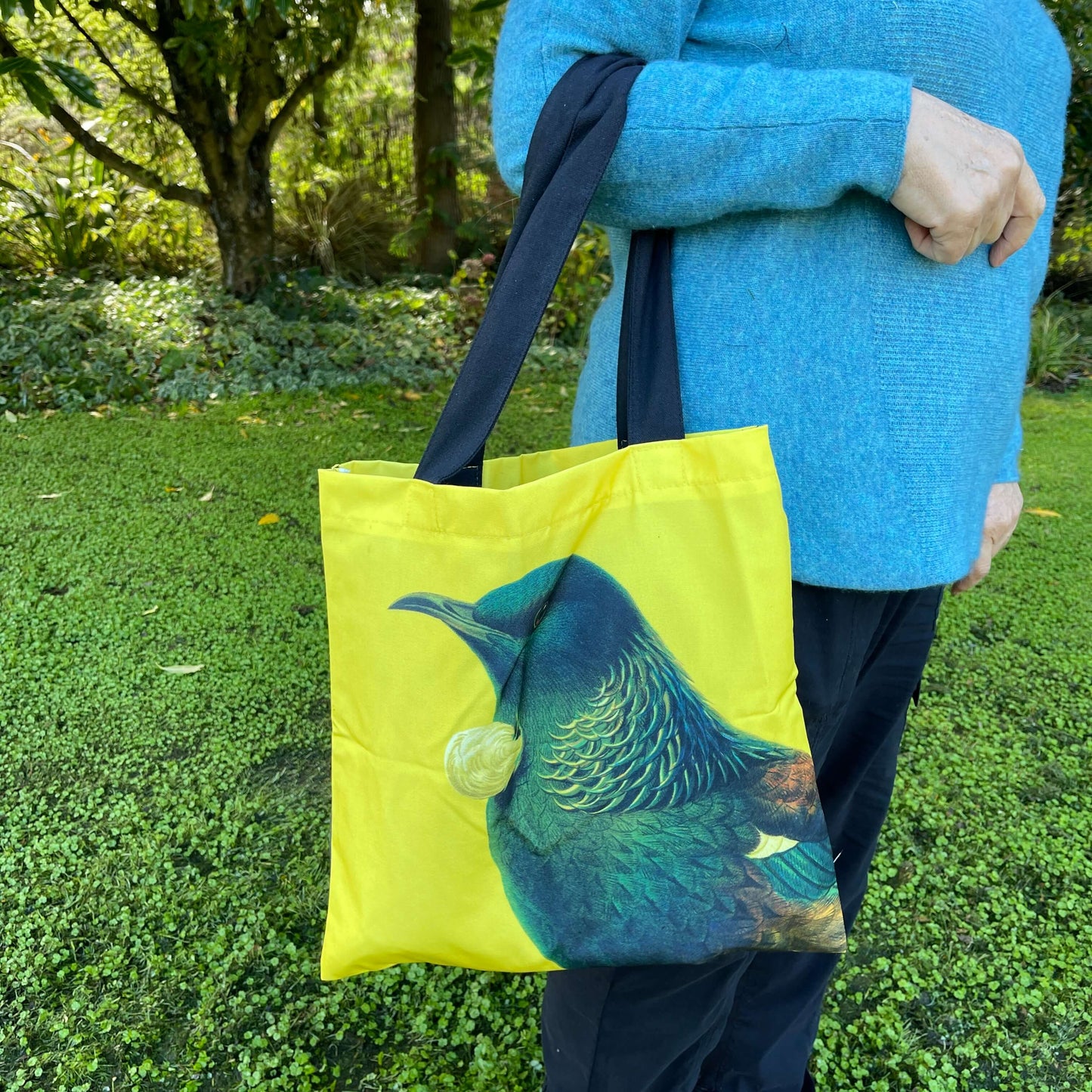 Woman with a tote bag over her arm. The bright yellow tote teatures a Tui bird.