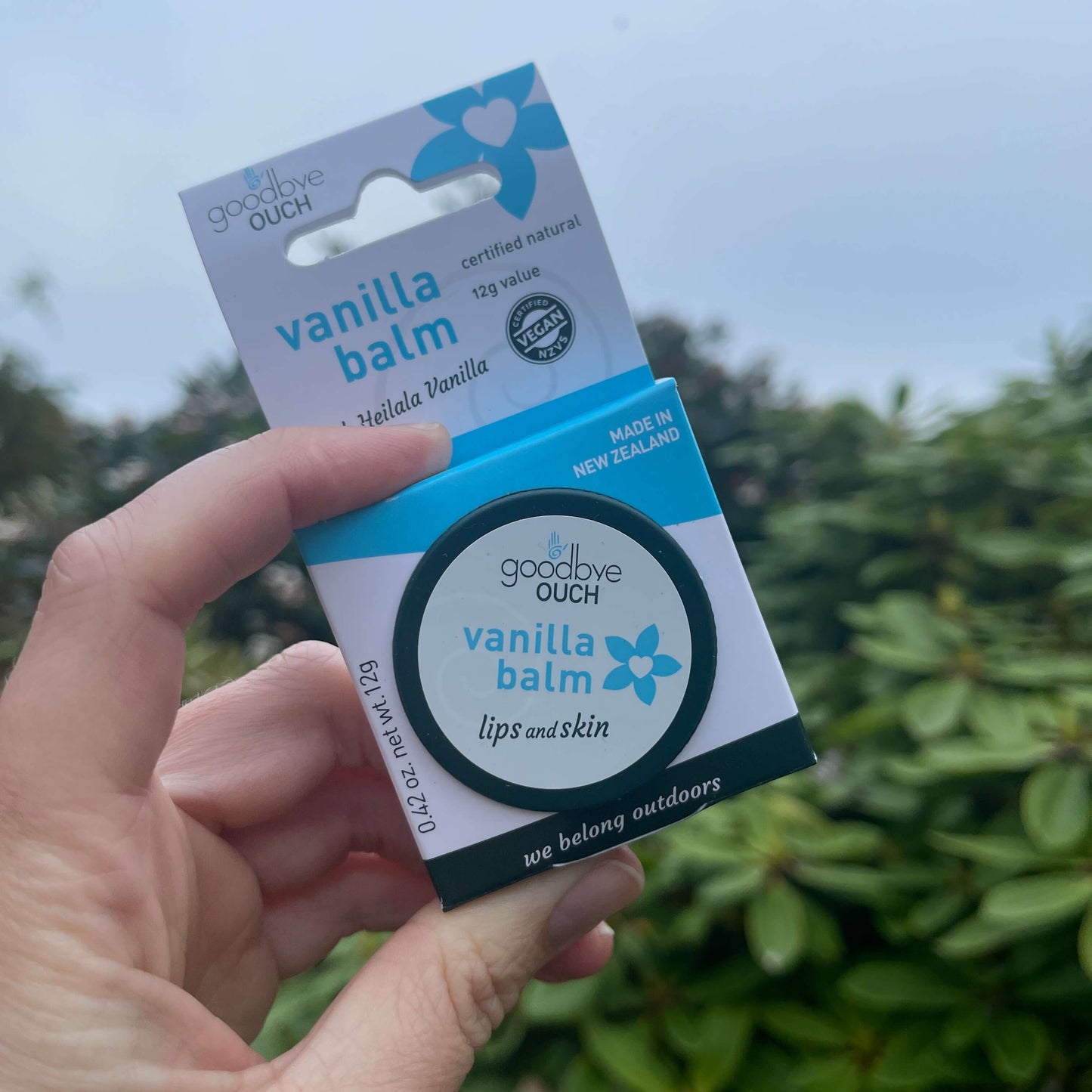 Persons hand holding a small tin of Vanilla Balm inside the cardboard packaging.
