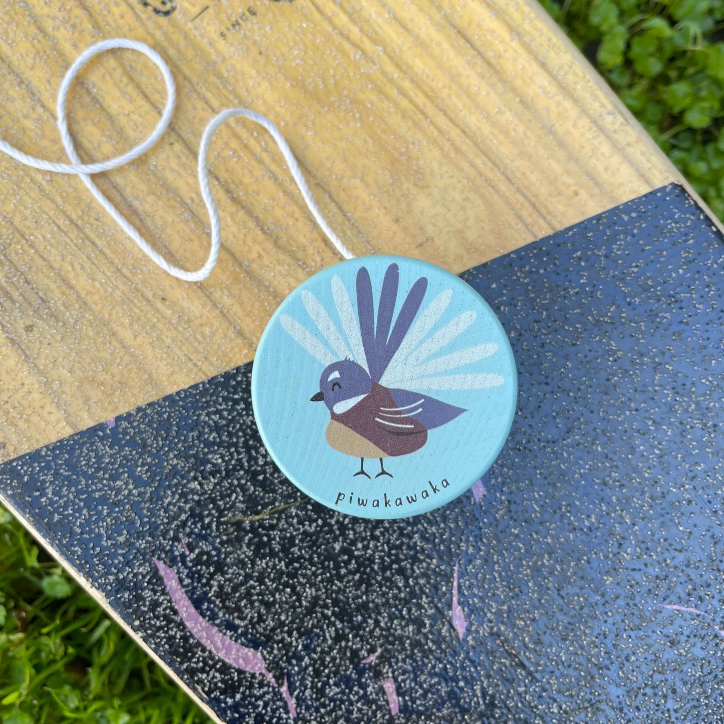 A sky blue wooden yoyo with a Fantail bird painted on it sitting on a skateboard.