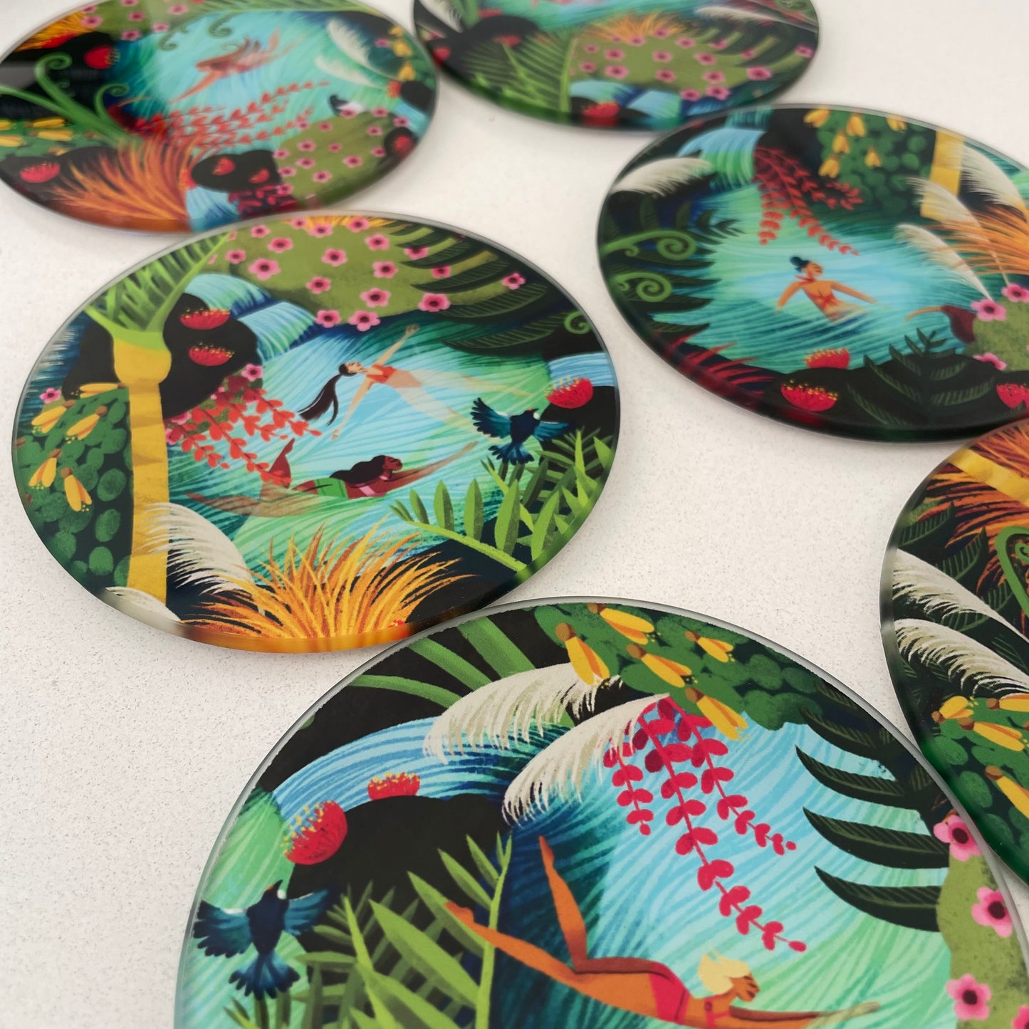 Close up image of 6 round glass coasters featuring bright colourful artwork of native New Zealand flora & fauna and women swimming in water.