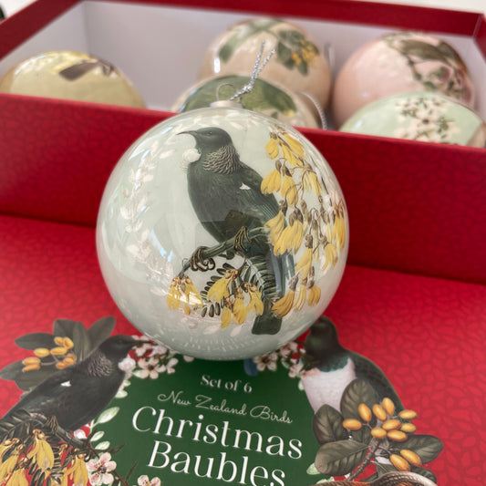 Pale green Christmas bauble with a Tui bird and kowai flowers on it. Placed on a red box lid with a red box in the background filled with 5 other baubles.