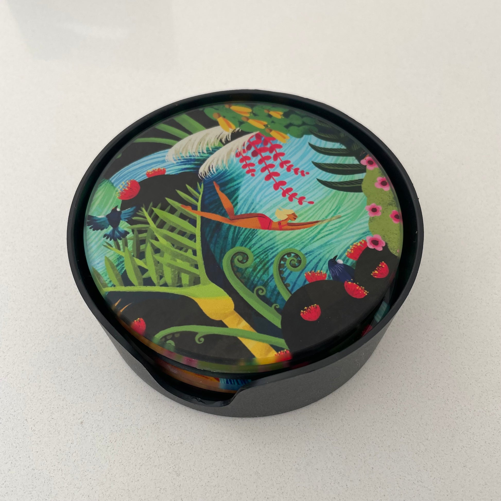 Set of 6 round glass coasters featuring bright colourful artwork of native New Zealand flora & fauna and women swimming in water. Placed in a black stacking container.