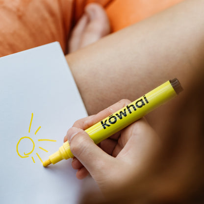 Person holding a yellow marker labeled 'kowhai' and drawing a sun on a piece of paper.