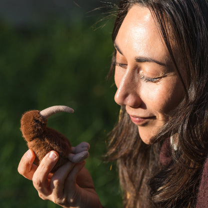 A woman outdoors holds a small brown plush Kiwi bird toy with a curved cream-colored beak in her hands, with a blurred green natural background.