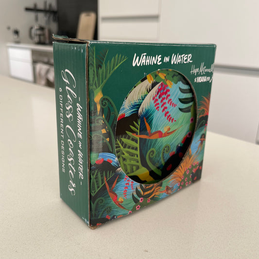 Dark green box with glass coasters inside sitting on a kitchen bench. The box and coasters also feature bright colourful artwork of native New Zealand flora & fauna and women swimming in water.