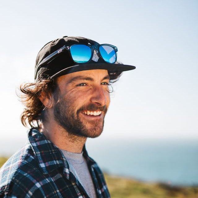 A smiling man outdoors wearing a black baseball cap with blue mirrored sunglasses on the brim, a dark plaid flannel shirt over a gray t-shirt, with a blurred coastal landscape in the background.