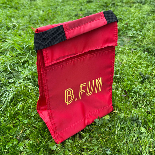Insulated lunch carry bag in red with yellow text saying B.FUN sitting on green grass.