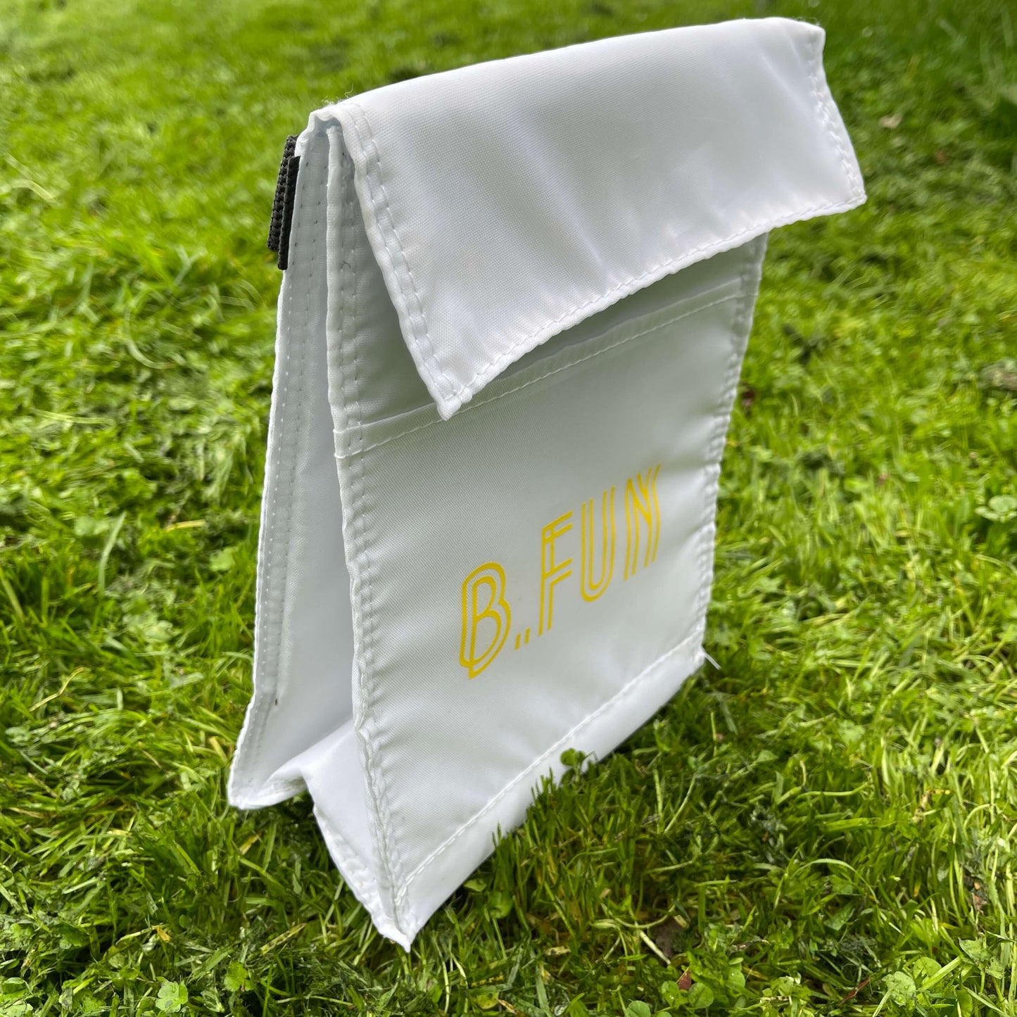 Insulated lunch carry bag in white with yellow text saying B.FUN sitting on green grass.