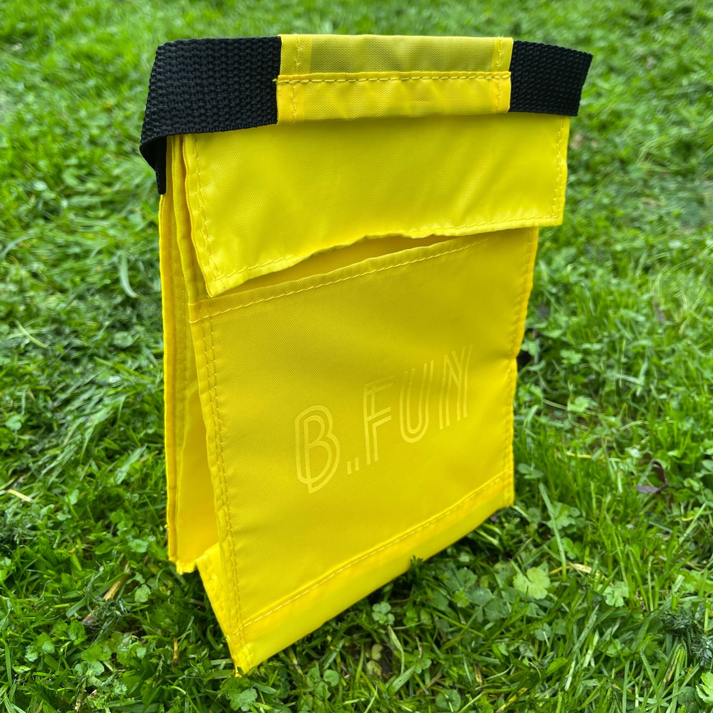 Insulated lunch carry bag in yellow with yellow text saying B.FUN sitting on green grass.