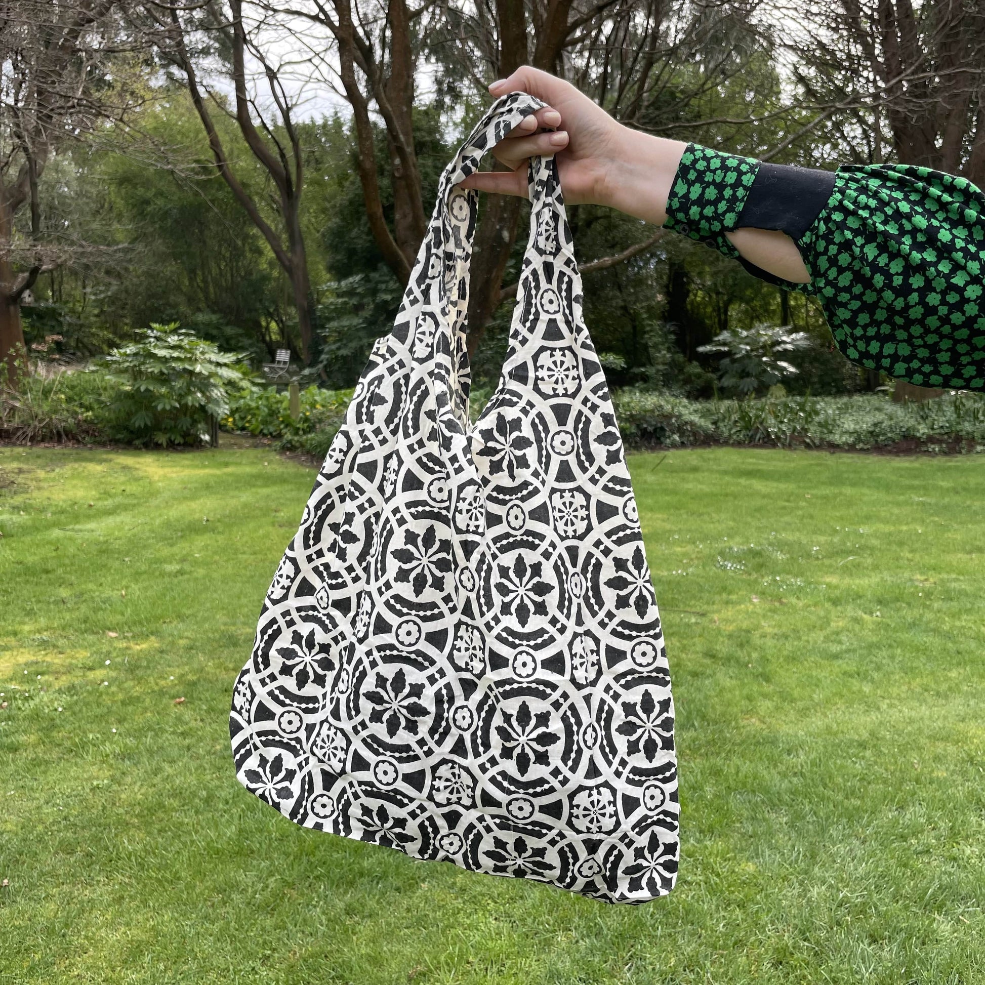 Womans hand holding a patterned black and white cotton tote bag with a lush green garden in the background.
