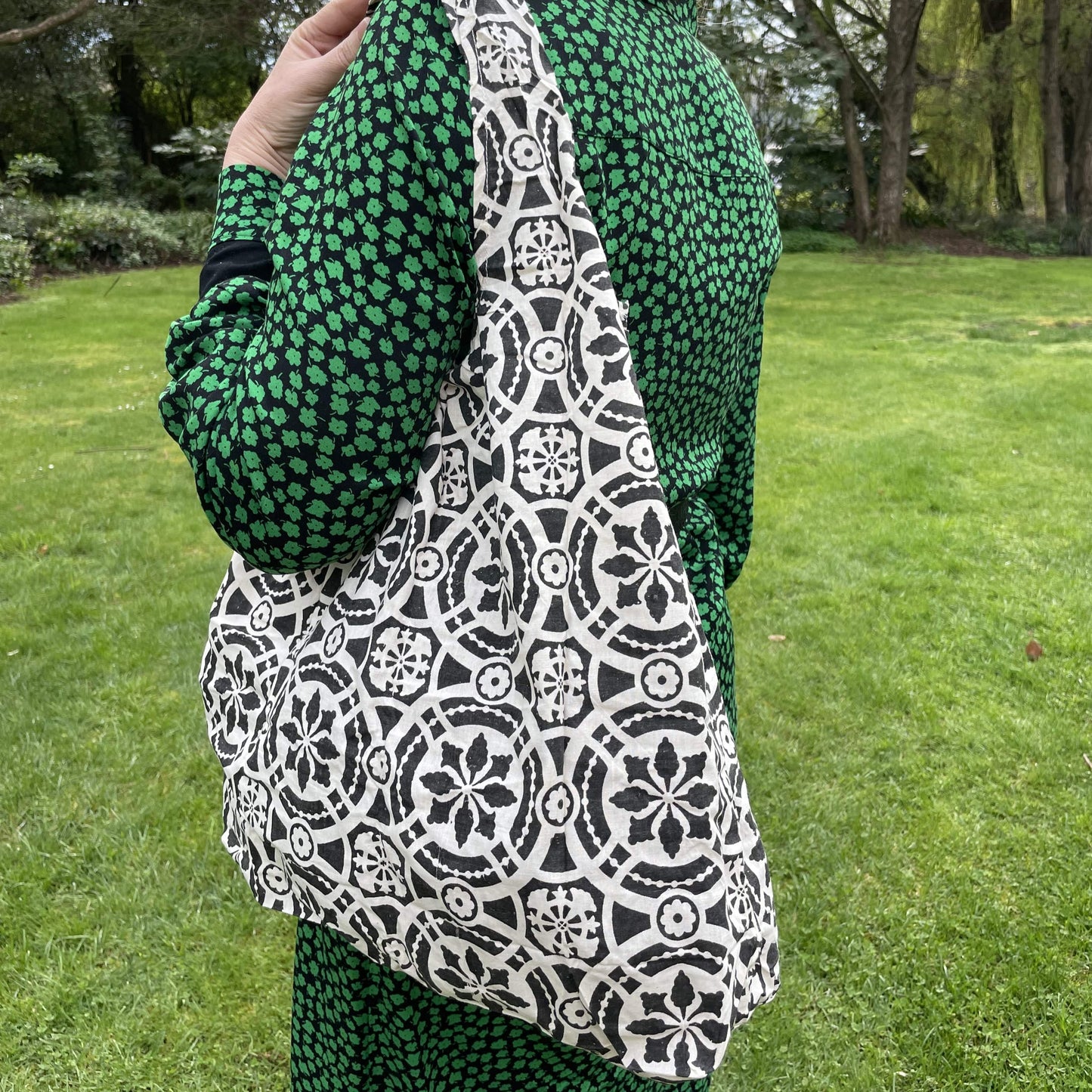 Close up of a woman wearing a black and green dress. She has a black and white patterned reusable cotton tote bag over her shoulder. A green garden setting is the background.