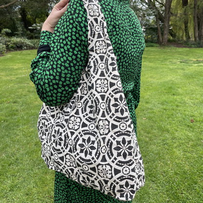 Close up of a woman wearing a black and green dress. She has a black and white patterned reusable cotton tote bag over her shoulder. A green garden setting is the background.