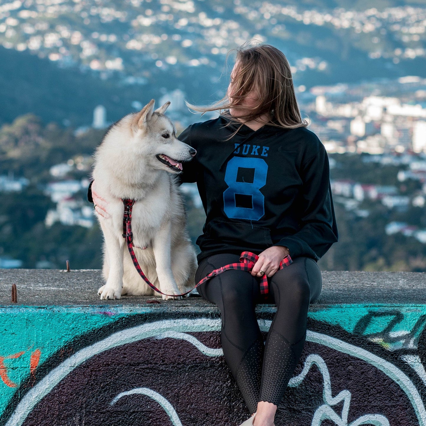 Woman and dog sitting on a wall. Woman is holding a red and black checked dog leash.