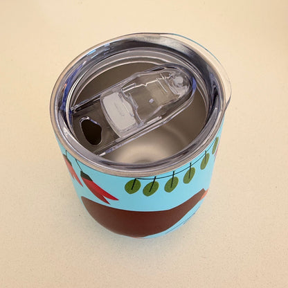 A close-up of a stainless steel travel tumbler with a clear lid and flip-up straw, decorated with a light blue background and brown kiwi bird design featuring red kaka flowers and green leaves.