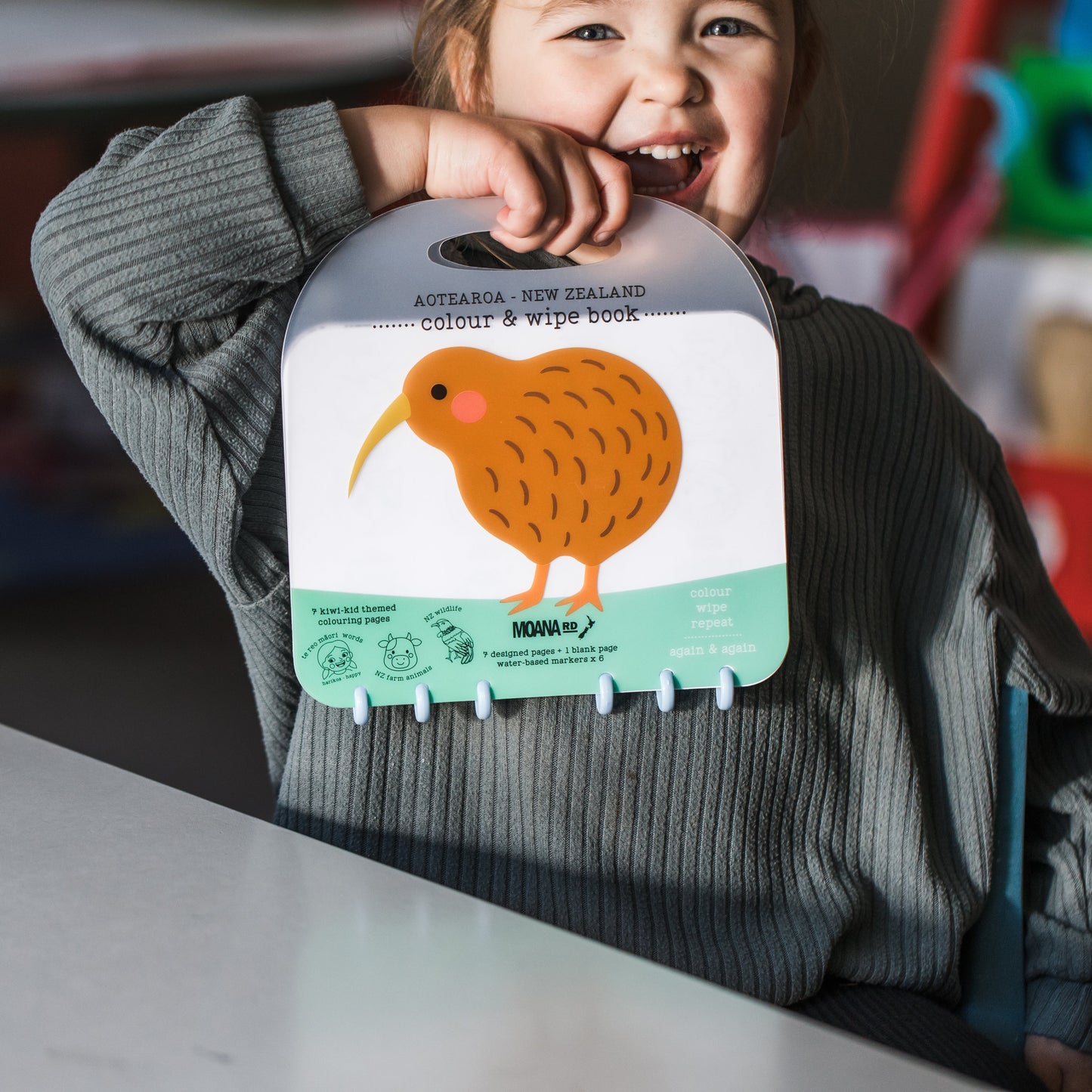 A smiling child in a gray ribbed sweater holds a Kiwi Books 'Colour & wipe book' featuring a kiwi bird illustration on the front, set against a softly blurred indoor background.