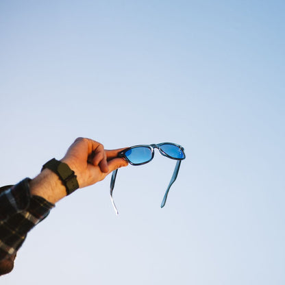 A hand wearing a black watch holds a pair of sunglasses with blue lenses and grey frames against a clear light blue sky.