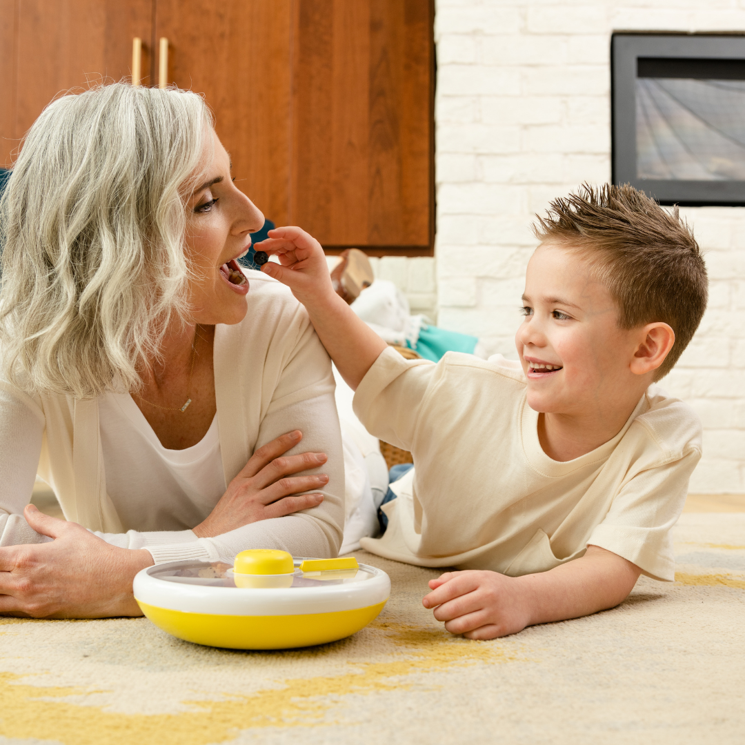 Child feeding mother from a Gobe snack spinner lunch box in lemon yellow and white.