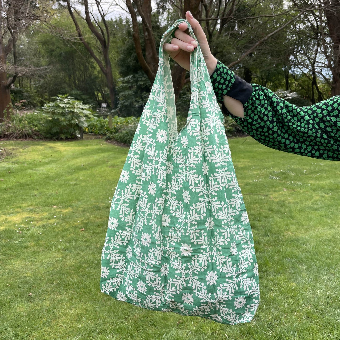 Womans hand holding a floral patterned green and white cotton tote bag with a lush green garden in the background.