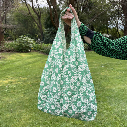 Womans hand holding a floral patterned green and white cotton tote bag with a lush green garden in the background.