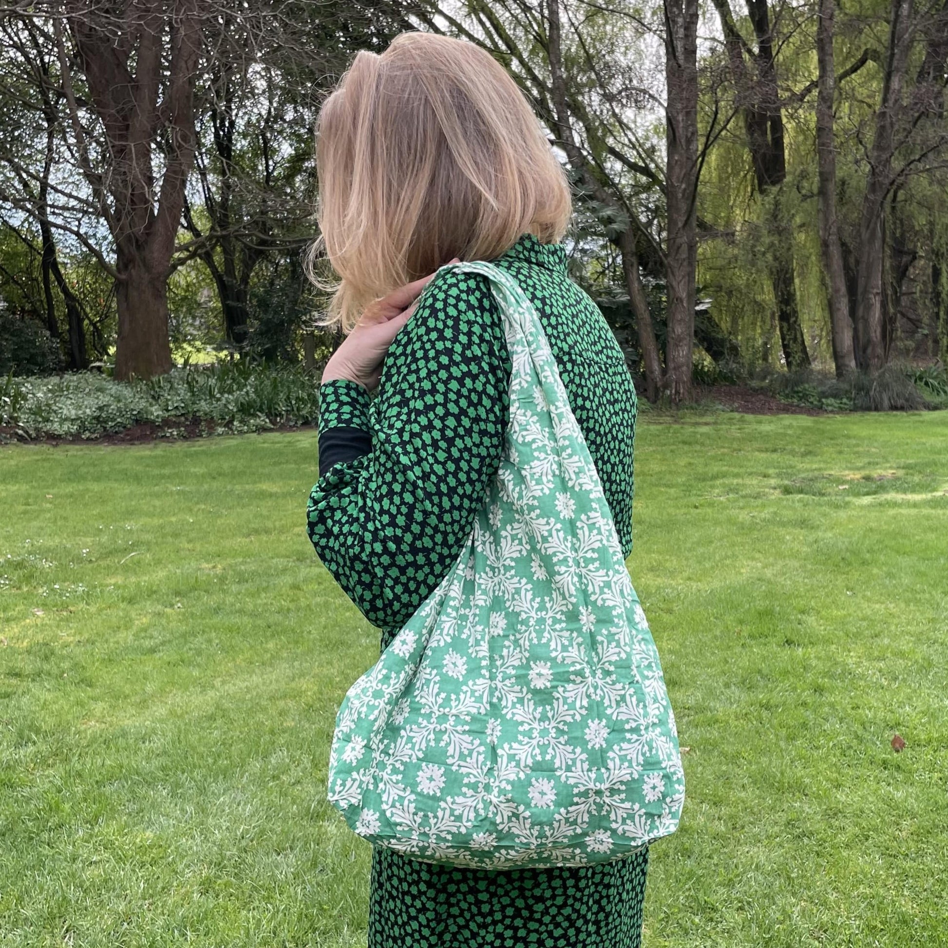 Woman standing in a garden in a black and green dress with her back to the camera. She has a green and white floral patterned reusable cotton tote bag over her shoulder.