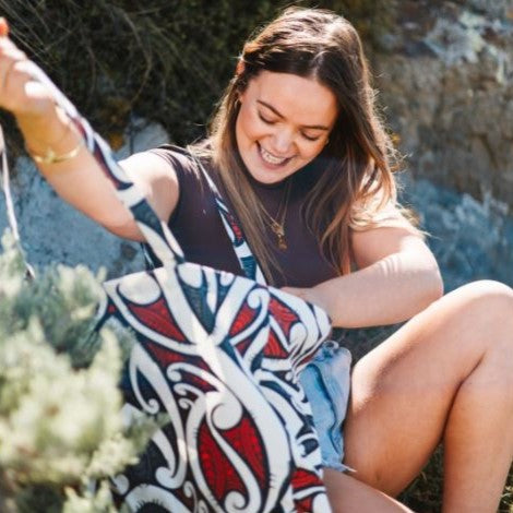 Woman sitting outdoors with a tote bag with red, black and white maori design.