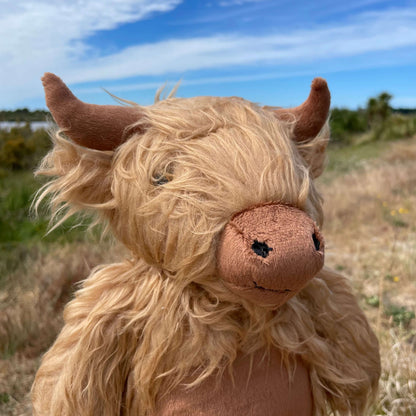Close up of a soft toy Highland Cow sitting on a fence post wiith an estuary view behind it. The toy has a shaggy light brown fur, darker brown horns, nose and tummy. 