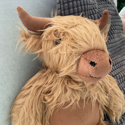 Close up of a soft toy Highland Cows face. It has a shaggy light brown fur, darker brown horns, nose and tummy. 