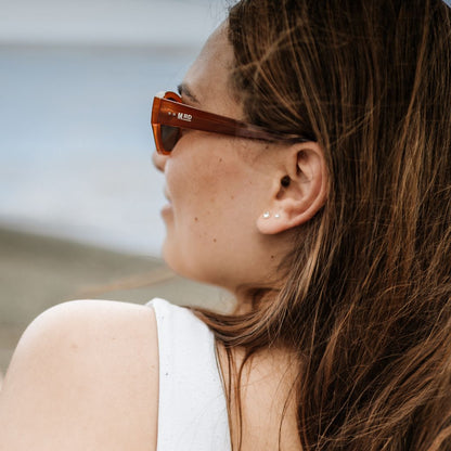 Close up of a womans face side on wearing burnt orange coloured plastic frame sunglasses with brown polarized lenses. A beach scene is out of focus in the background.