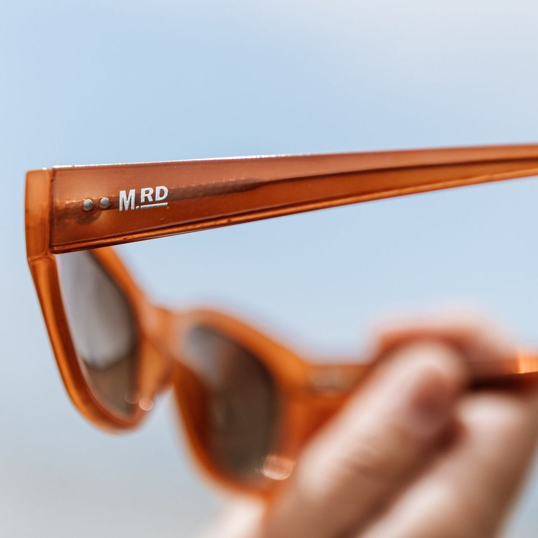 Burnt orange coloured plastic frame sunglasses with brown polarized lenses being held in someones hand. Mostly out of focus except for the sunglasses arm which shows the branding of Moana RD.