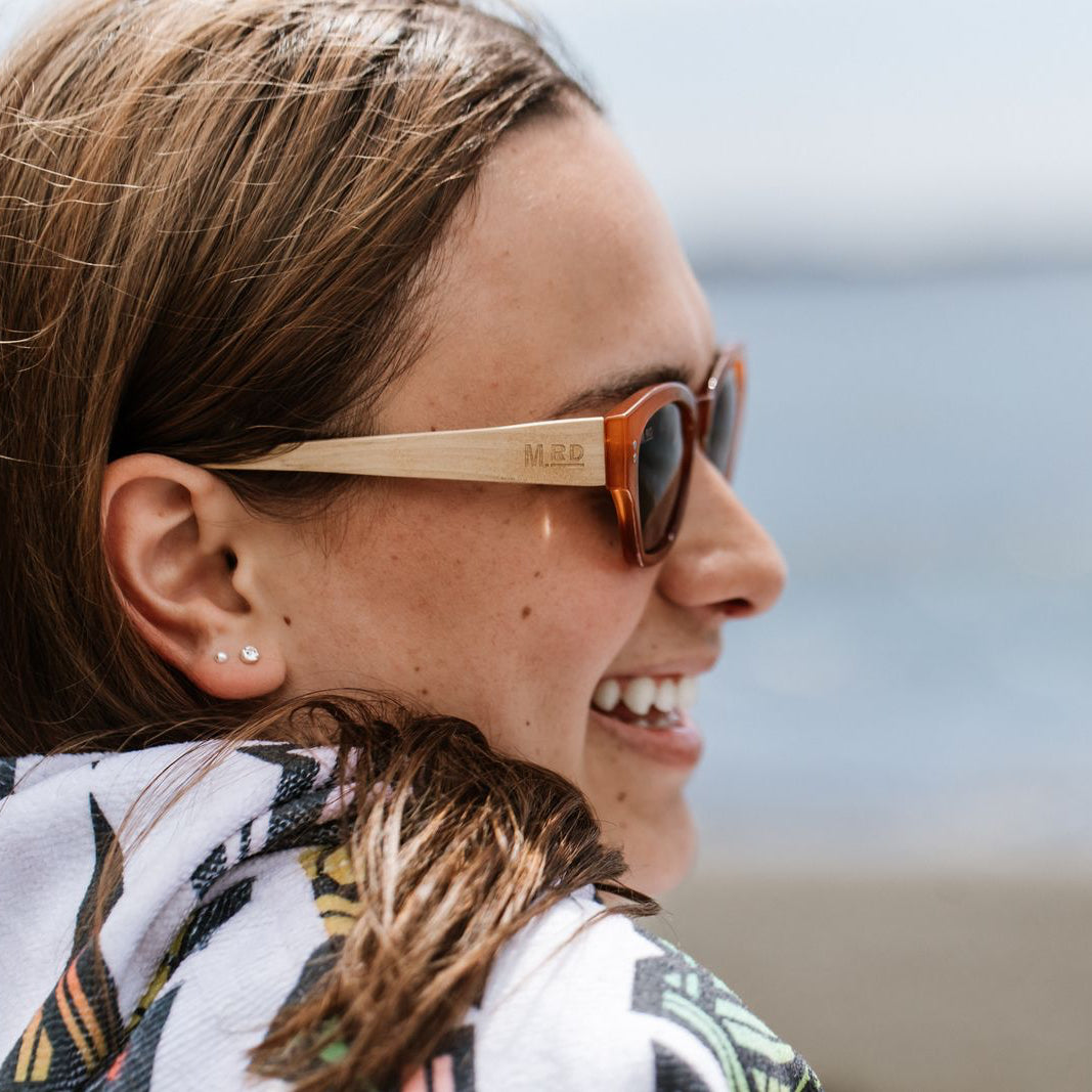 Close up of a womans face side on wearing burnt orange coloured plastic frame sunglasses with brown polarized lenses and wooden arms. A beach scene is out of focus in the background.